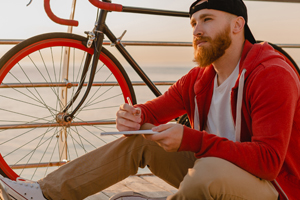 Man sitting on a bridge next to his bike writing in a journal