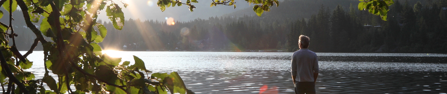 Man standing on the lakeshore looking over a forest into the sun
