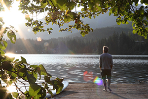 Man standing on the lakeshore looking over a forest into the sun