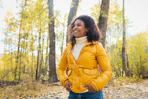Woman with a white turtle neck and yellow coat smiling in the woods