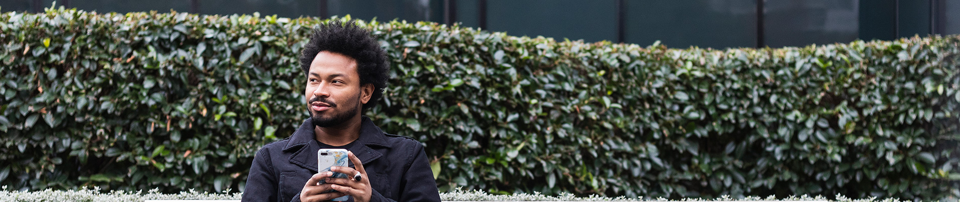 A young man standing by a hedge holding a cell phone