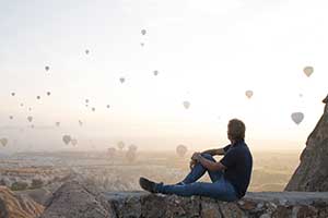 A man sitting on a rock plateau watching hot air balloons take to flight