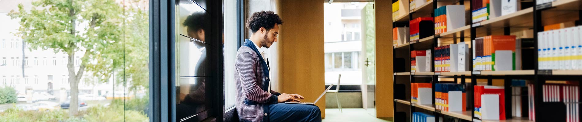 A young man sitting on a bench in a library like room typing on a lap top