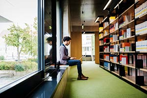 A young man sitting on a bench in a library like room typing on a lap top