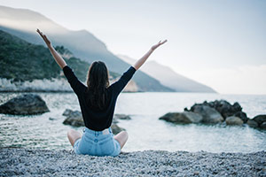 A woman sitting on a sandy area above a large body of water with arms stretched up to the sky
