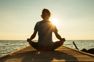 man sitting on a dock in a yoga type position facing a sun rise
