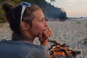 a young woman sitting by a small fire in the sand along side the ocean in thought
