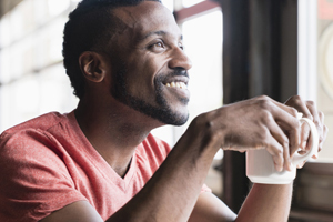 a young man sitting holding a cup of coffee looking out a window smiling
