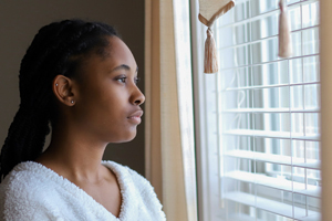 a young woman looking through the window shades to a bright outside