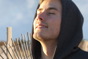 A man in a hoody, head tilted up into the sunlight next to a fence along sand dunes